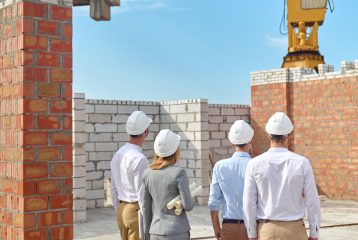 Back view of a supervisor with technical drawings and builders standing on the construction site